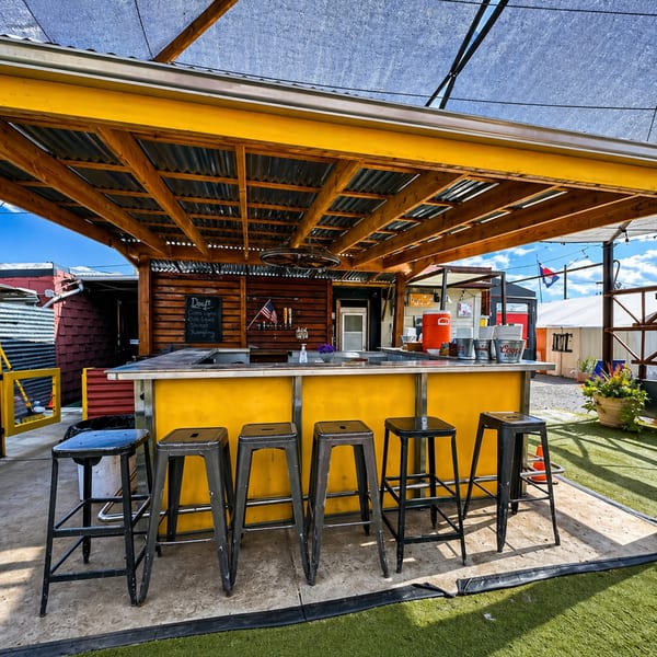 Outdoor bar with a yellow counter, black stools, wooden ceiling, and a shaded patio area.