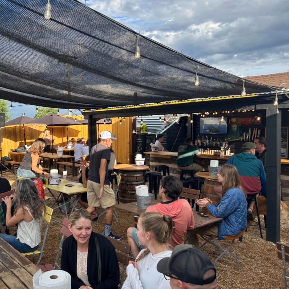 Busy outdoor restaurant patio with communal tables, patrons, string lights, shade canopy, and a full bar.