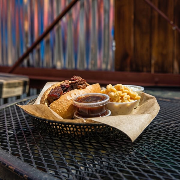 A BBQ brisket sandwich, mac and cheese, and dipping sauces in a basket on a metal table.