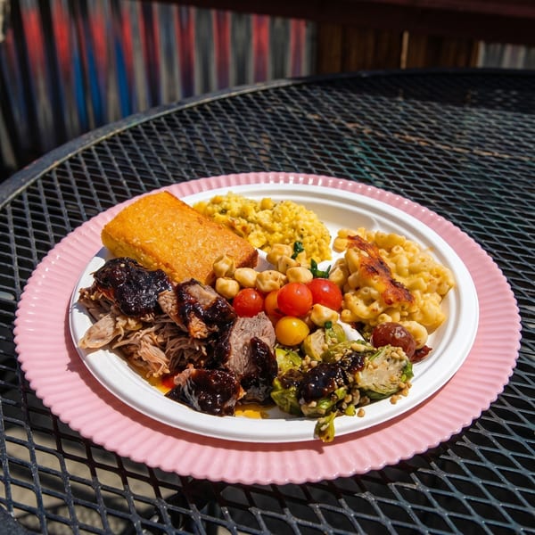 A BBQ plate with pulled pork, brisket, cornbread, mac and cheese, corn, tomatoes, and Brussels sprouts.