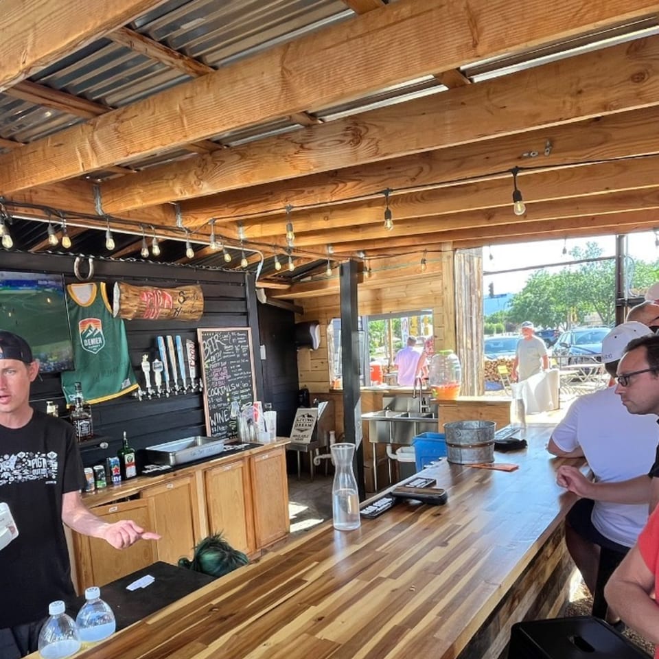 Rustic open-air bar with wooden beams, string lights, beer taps, chalkboard menu, bartender, and patrons seated at a long counter.
