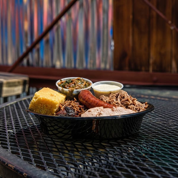 A black basket of barbecue with pulled pork, sausage, cornbread, collard greens, and sauce.