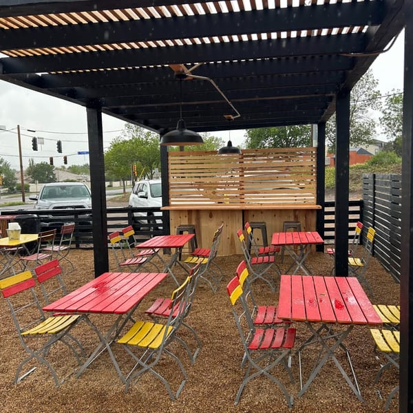 Covered restaurant patio with colorful red and yellow tables, folding chairs, hanging lamps, and a wooden privacy partition.