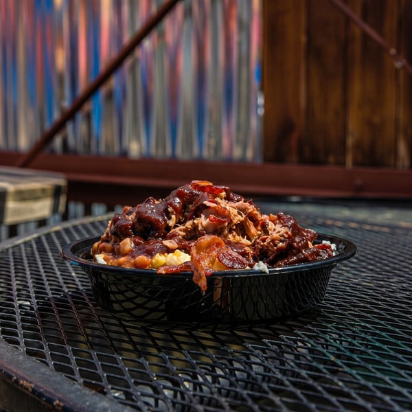A black bowl filled with barbecue pulled pork, beans, and mac and cheese on a metal table.