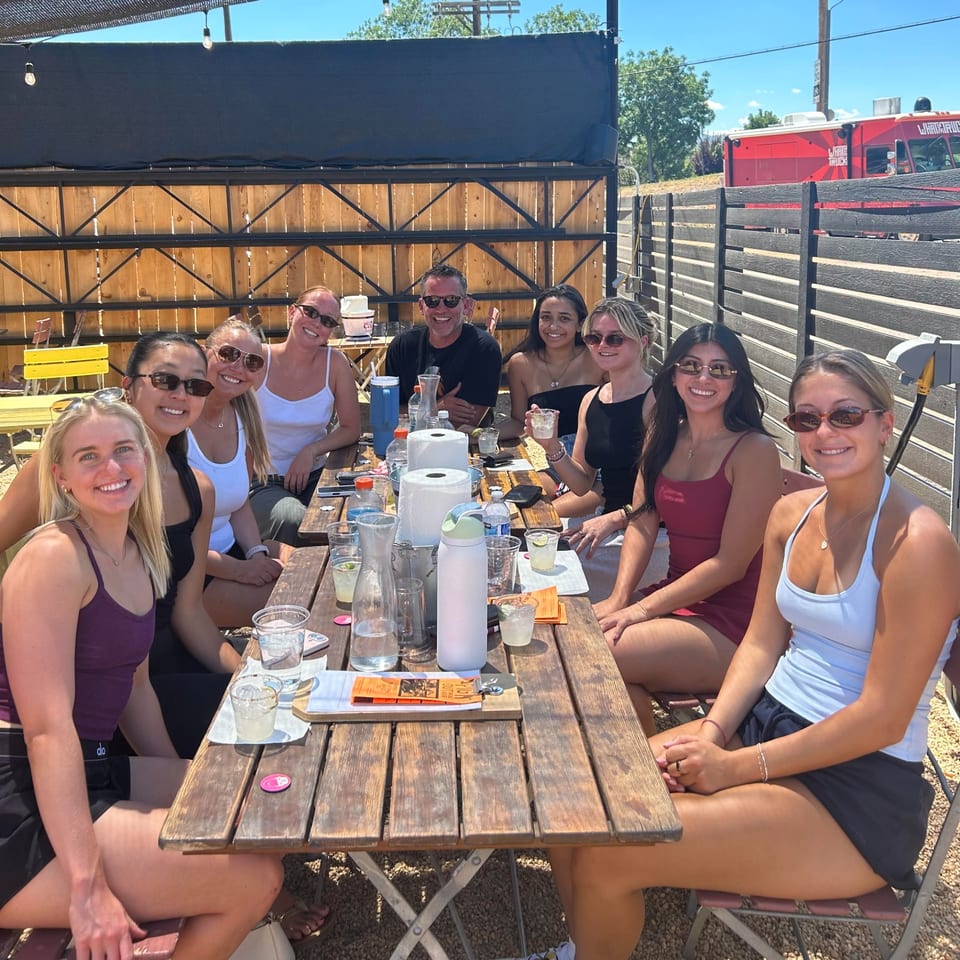 Group of friends smiling around a picnic table on a sunny outdoor patio, with drinks and a red food truck in the background.