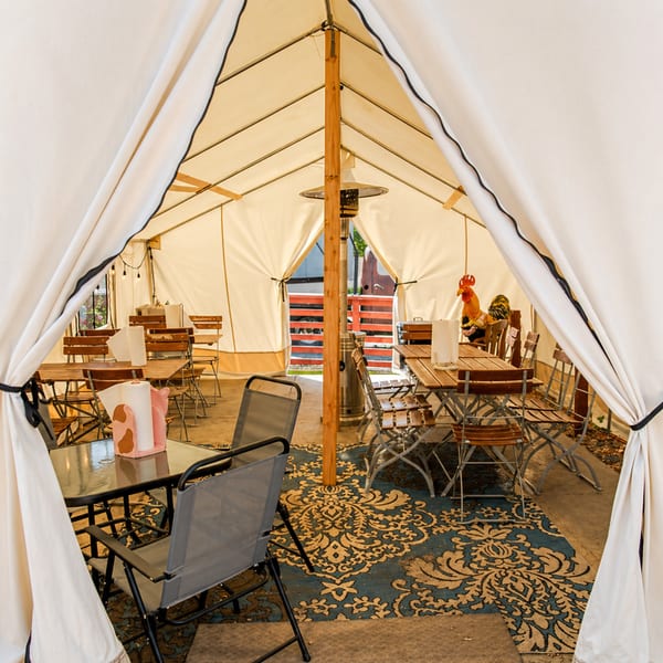 Outdoor dining tent with wooden tables, metal chairs, a patterned rug, and a rooster statue.