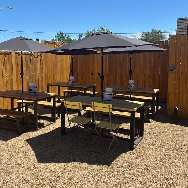Outdoor restaurant patio with wooden picnic tables, black umbrellas, benches, and folding chairs on a gravel surface.