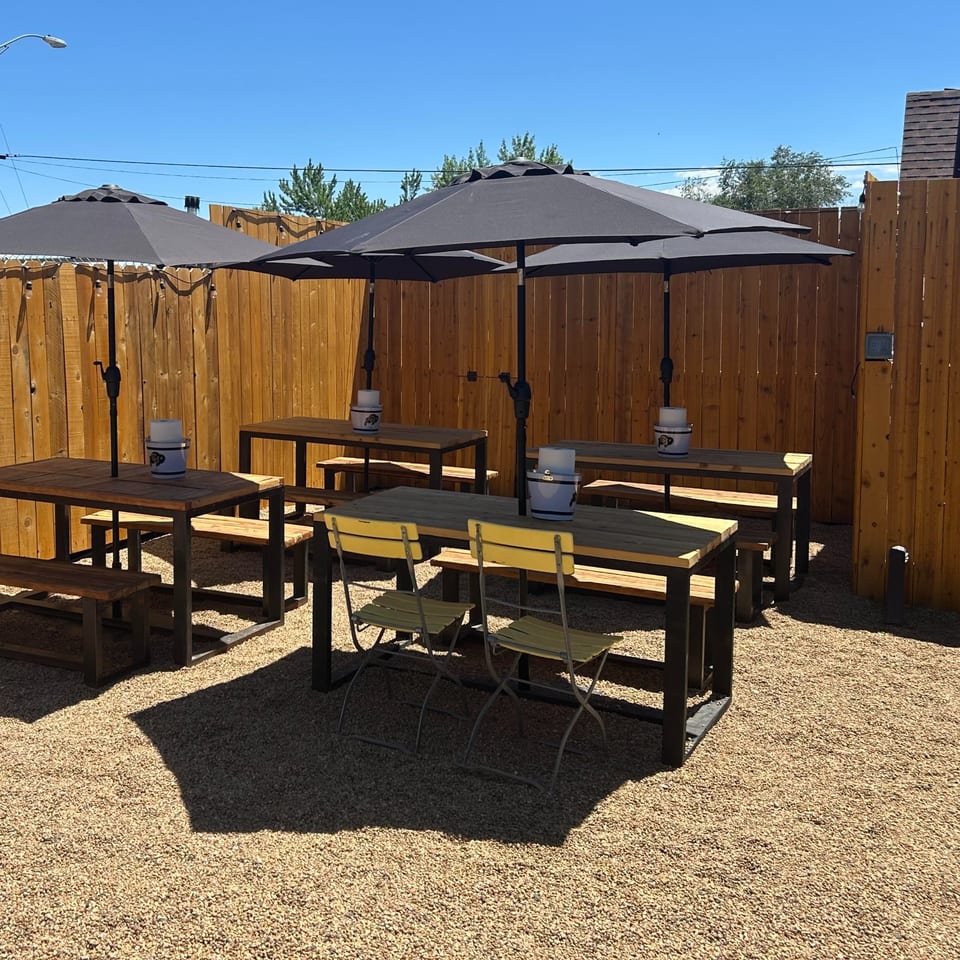 Outdoor restaurant patio with wooden picnic tables, black umbrellas, benches, and folding chairs on a gravel surface.