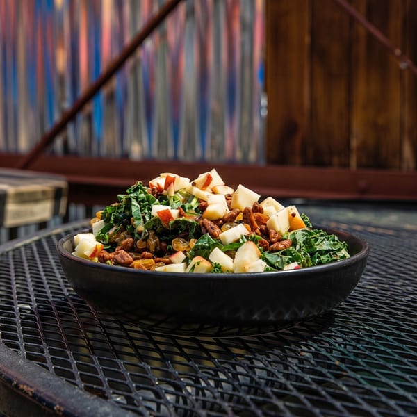 A black bowl of kale salad with apples, pecans, and golden raisins on a metal grate table.