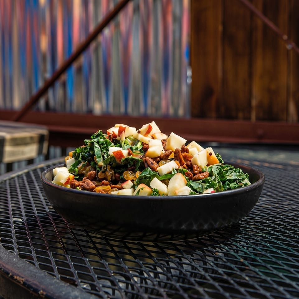 A black bowl of kale salad with apples, pecans, and golden raisins on a metal grate table.