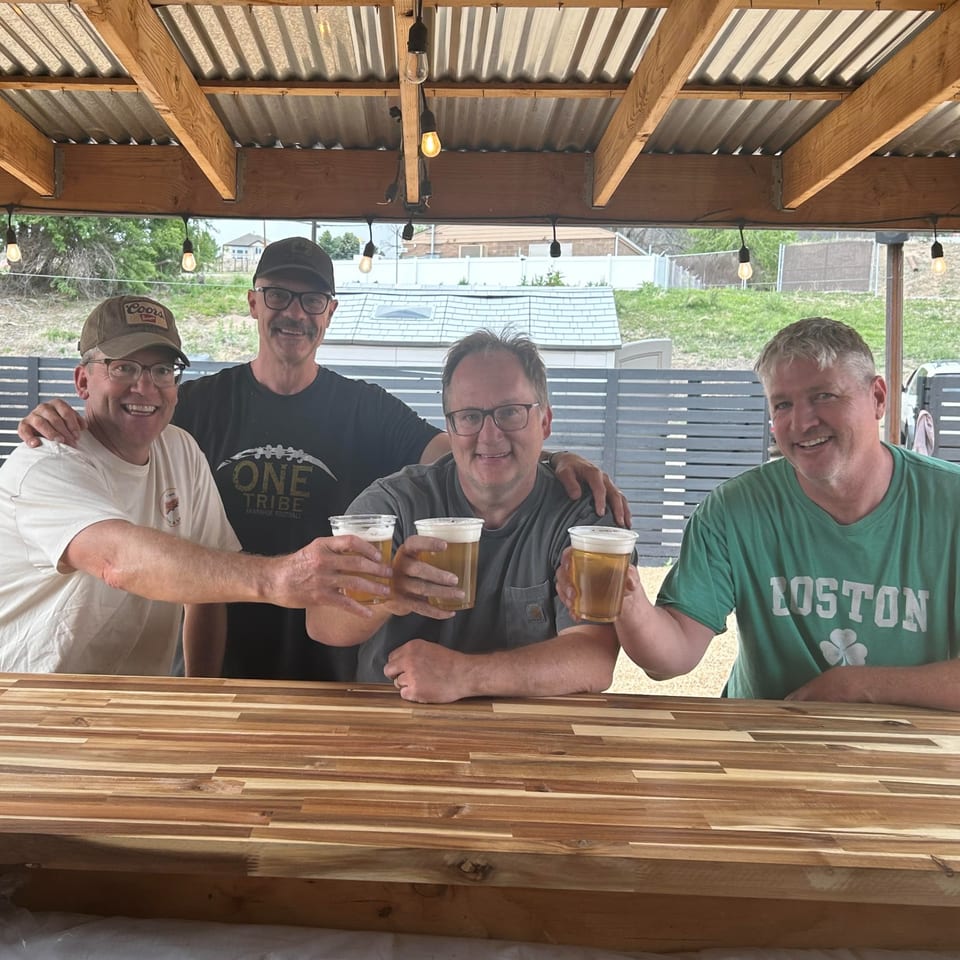Four men smile and raise plastic cups of beer at a covered outdoor bar with wooden counters and string lights.