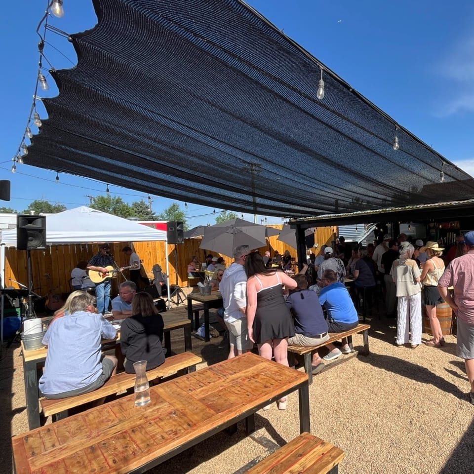 Outdoor restaurant patio with wooden picnic tables, string lights, a live acoustic guitarist, and a lively crowd beneath shade sails.