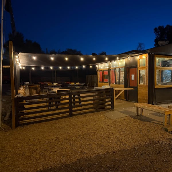 Outdoor restaurant patio with wooden tables, string lights, gravel ground, and a warmly lit entrance at dusk.