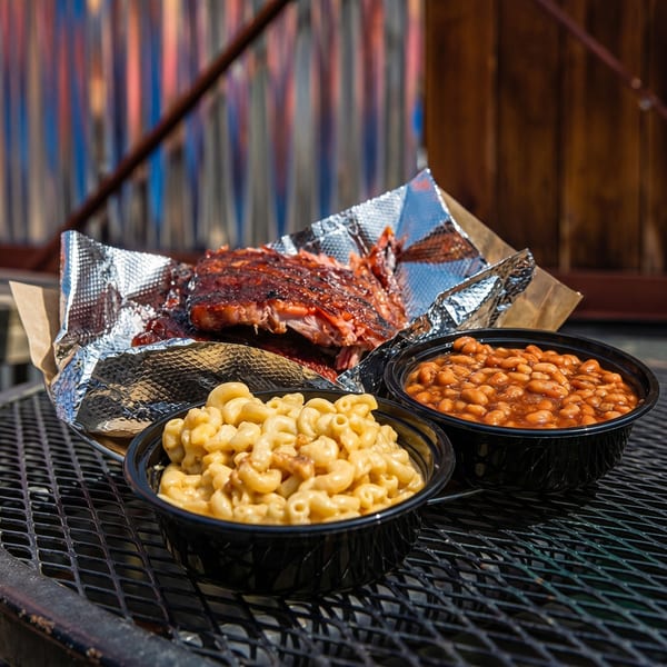 BBQ ribs in foil with sides of mac and cheese and baked beans on a metal table.